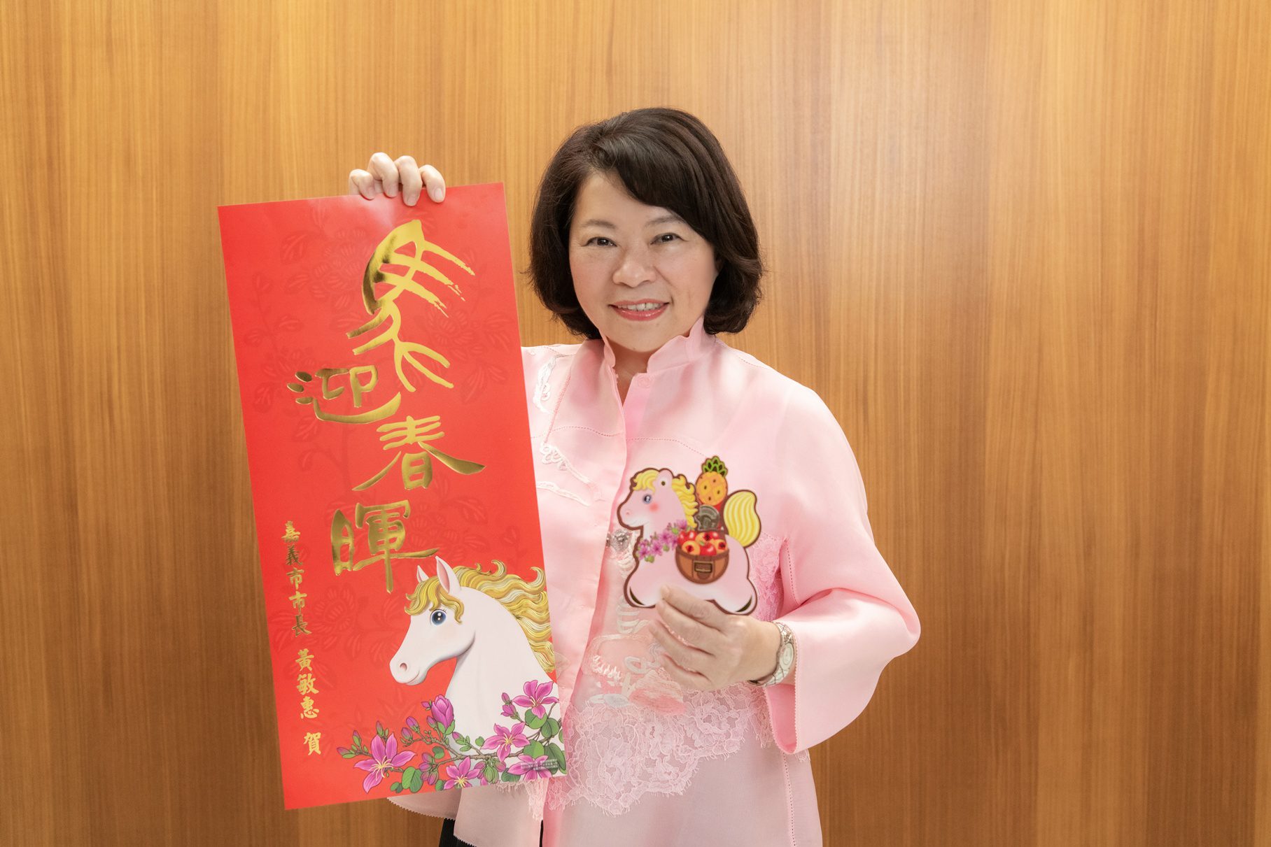 ▲市長黃敏惠開心展示分別由插畫家、書法家獨特設計的「粉紅超馬」小紅包以及「馬迎春暉」春聯。(圖/嘉義市政府提供)